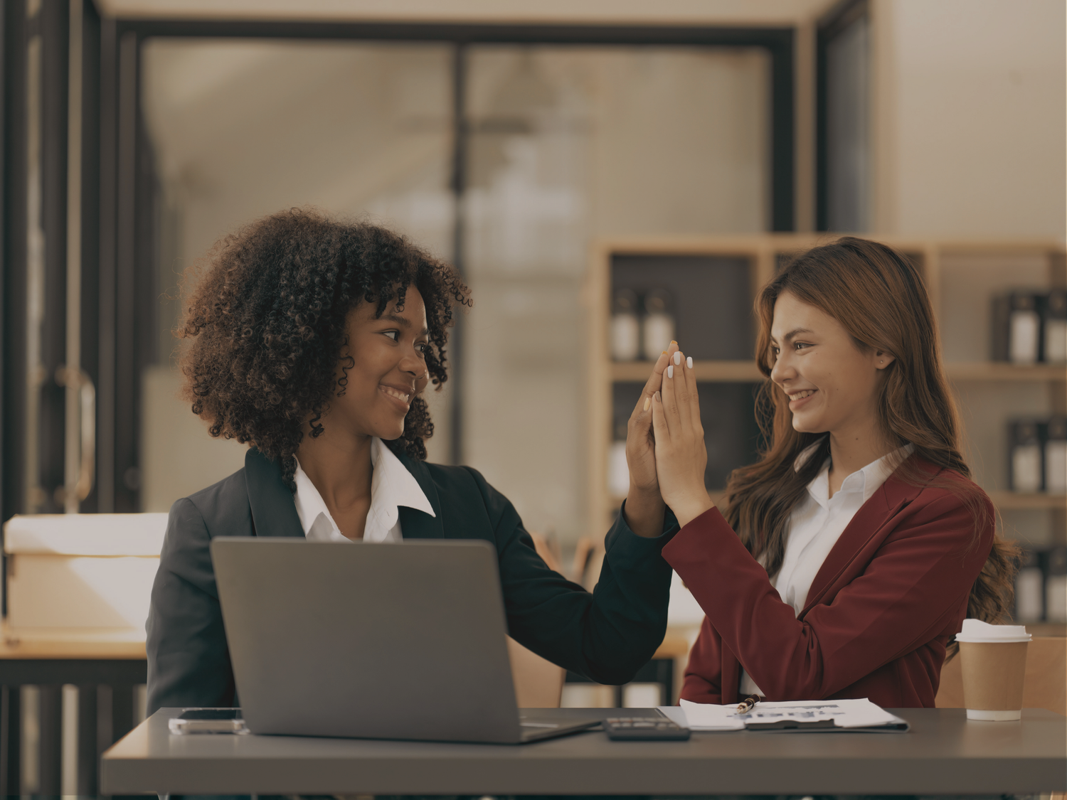 Two women high-fiving at a desk with a laptop and coffee cup in an office setting.