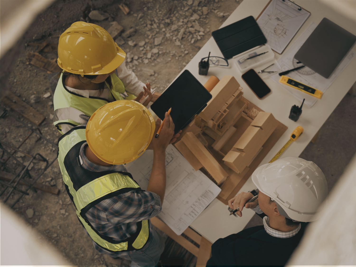 Three construction workers in hard hats and safety vests discussing plans at a table with tools and documents.